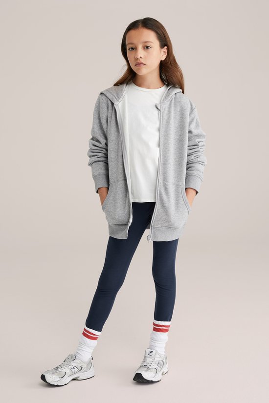 Meisje in donkerblauwe legging, wit t-shirt en grijze hoodie, staand.