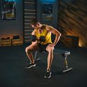 Een man in een gele tanktop en zwarte shorts doet een dumbbell-oefening op een zwarte trainingsbank met 'TX 100B' op de zijkant.