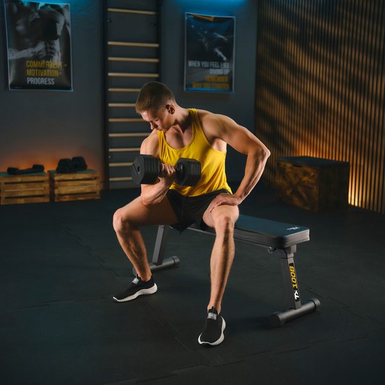 Een man in een gele tanktop en zwarte shorts doet een dumbbell-oefening op een zwarte trainingsbank met 'TX 100B' op de zijkant.