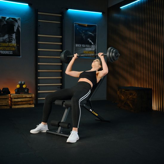 Vrouw met zwarte sportkleding ligt op een zwarte fitnessbank en tilt een halterstang omhoog.