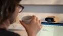 Vrouw met bril drinkt koffie bij een laptop met webcam.