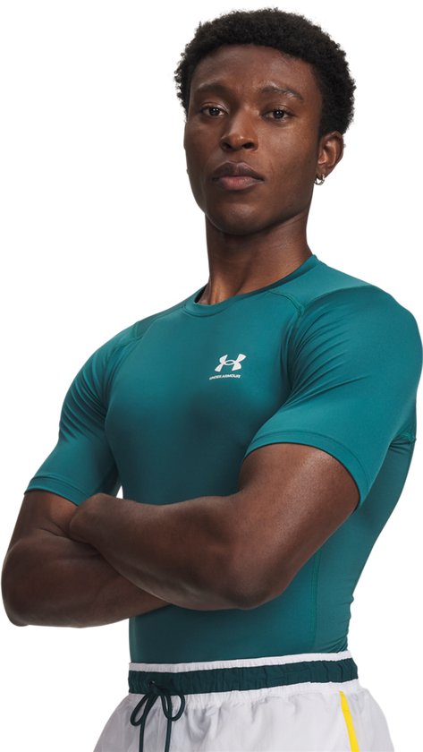 Man met gekruiste armen in groen Under Armour sportshirt. Witte sportbroek met gele strepen.