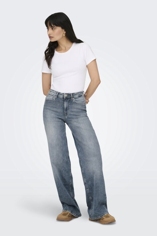 Model in een witte top en wijde, lichtblauwe spijkerbroek. Ze draagt bruine schoenen en staat met de handen achter de rug.