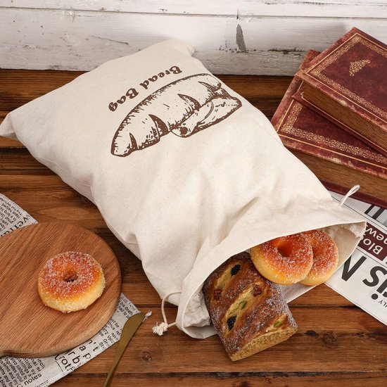 Off-white canvas zak met broodprint, gevuld met brood en donuts. Naast boeken en een houten plank.