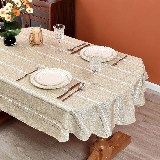 Nappe ovale aspect lin, rayures blanches verticales et décorations brodées. Dressée avec vaisselle, fleurs et chaises.