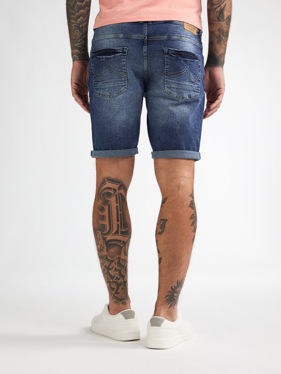 Man, staand, met de rug naar de camera, in een roze T-shirt en spijkerbroek met opgerolde zoom, witte sneakers.