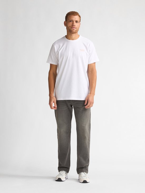 Man met rood haar, gekleed in een wit T-shirt met oranje logo op de borst en grijze jeans.