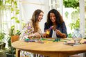 Twee vrouwen, zittend aan een ronde tafel, bouwen aan LEGO bloemen, met kleurrijke bloemen in paars, blauw en geel.