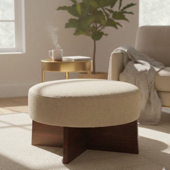 Pouf bouclé crème avec un pied croisé en bois marron foncé, dans un salon près d'un fauteuil.