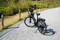 Zwarte elektrische fiets met bagagedrager op een grindpad, met draad-en-paal-afscheiding op de achtergrond.