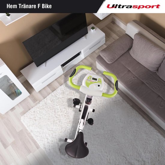Opvouwbare witte en groene hometrainer in een woonkamer, geplaatst op een wit vloerkleed bij een bank en tv-meubel.