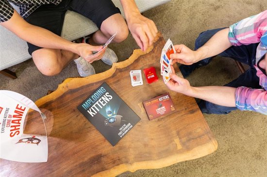 Vijf spelers spelen 'Exploding Kittens'. De tafel toont het basisspel, de uitbreidingsdoos, kaarten en een 'Cone of Shame'.