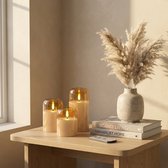 Drie matglazen LED-kaarsen met amberkleurige glazen kap en een afstandsbediening op een houten kastje, met boeken en pampasgras.