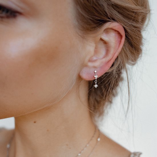 Une femme porte des boucles d'oreilles argentées avec trois petites zircones, suspendues à une chaîne.