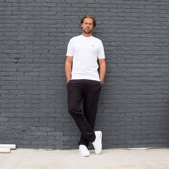 Man in wit Donnay t-shirt, zwarte broek en witte sneakers, leunt tegen donkergrijze bakstenen muur.