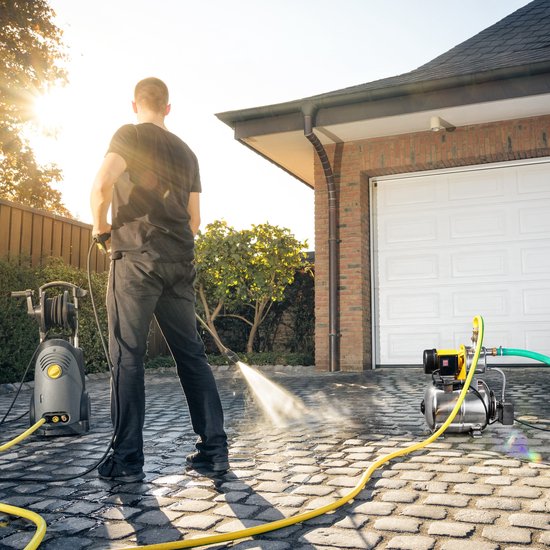 Een man met een hogedrukreiniger, terwijl een hydrofoorpomp met gele motorbehuizing op de oprit staat.