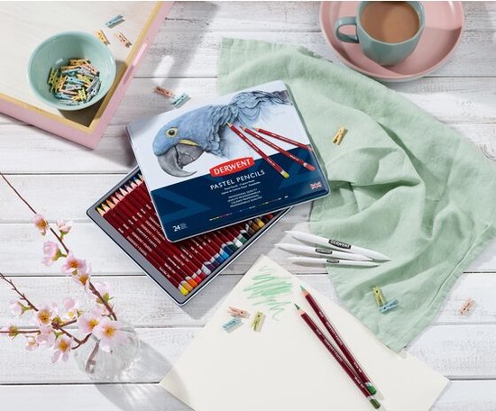 Een blik Derwent Pastel potloden en een open blik met potloden, naast een mok thee en een schetsboek.