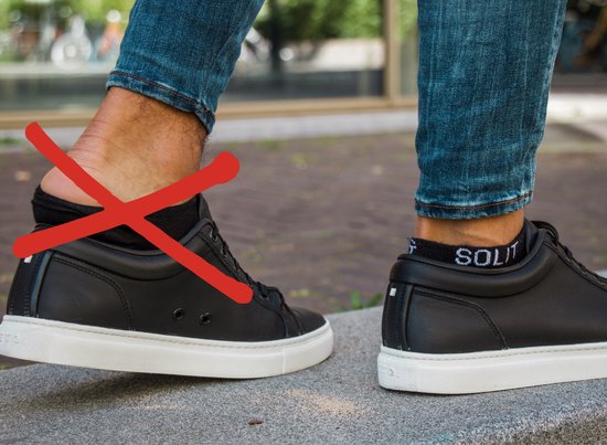 Zwarte lage sneakers met witte zolen, gedragen met een spijkerbroek. Rode kruis door een zichtbare enkelsok.