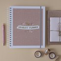 Een koraalkleurige ringband-planner met 'Myn Bruiloft Planner' ligt naast een glitter notitieboekje en een pen.