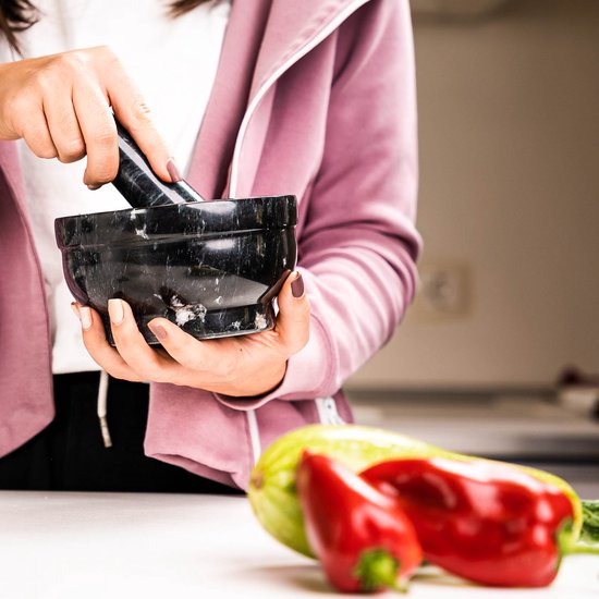 Vrouw in roze trui met zwarte marmeren vijzel en stamper, met rode peper en courgette op de voorgrond.