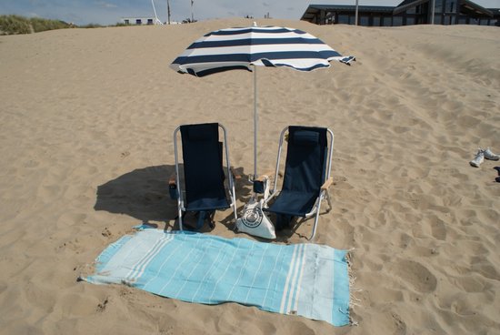 Twee donkerblauwe campingstoelen onder een gestreepte parasol op het strand. Een blauwwitte handdoek ligt ervoor.
