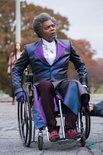 Samuel L. Jackson in een paars-blauwe glimmende jas zit in een rolstoel, met een witte das en paarse schoenen.