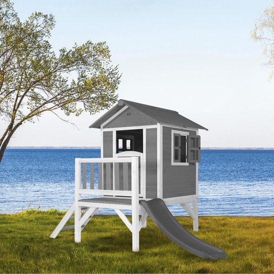 Grijs houten speelhuis met witte randen, veranda, trap, schuif en zichtbare ramen.