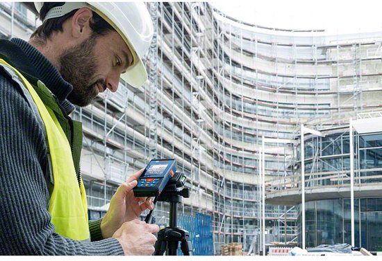 Man met veiligheidshelm en gele hes die een Bosch laser afstandsmeter vasthoudt op een bouwplaats.