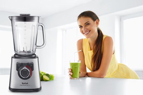 Vrolijke vrouw met een glas groene smoothie naast de Tefal Perfectmix+ blender in RVS.