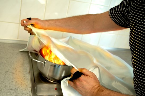 Handen die een witte brandblusdeken over een pan met vlammen leggen.