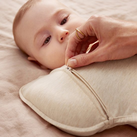 Een baby met bruine ogen wordt voorzichtig in een crèmekleurige slaapzak met ritssluiting gelegd.