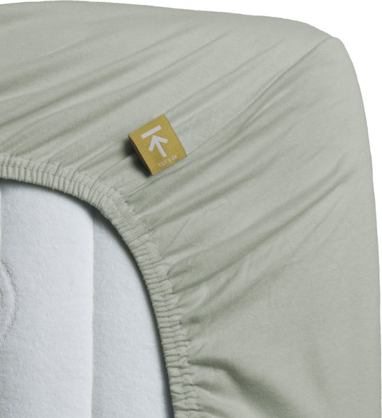 Drap-housse gris-vert en jersey pour surmatelas avec élastique et une étiquette jaune avec une flèche et 'top side'.