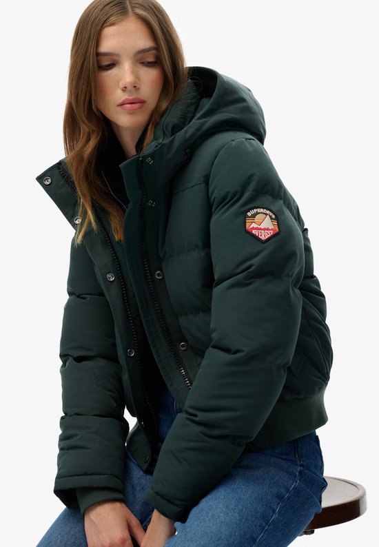 Vrouw in een donkergroene gewatteerde jas met capuchon, met een badge op de mouw en gedetailleerde naden.