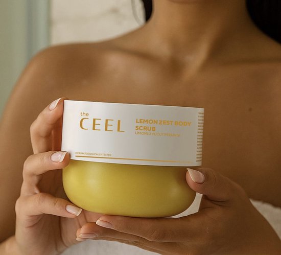 Een persoon houdt een geel potje 'Lemon Zest Body Scrub' van The Ceel vast.