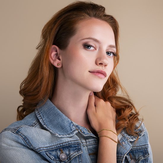 Model met rood haar draagt een gouden knopoorbel en een gouden schakelarmband, met spijkerjasje.