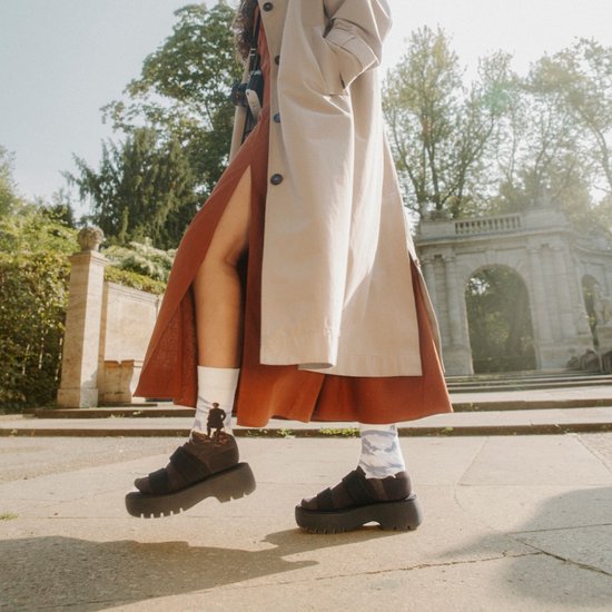 Model draagt een beige trenchcoat met een bruine split rok, witte sokken met een berglandschap en plateau sandalen.