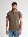 Man in een olijfgroen poloshirt en blauwe jeans, staand tegen een witte achtergrond.