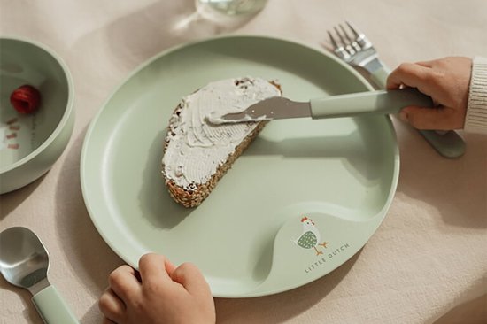 Assiette verte pour enfant avec image de poule, enfant tartine du beurre sur du pain avec un couteau. Doigts d'enfant visibles.