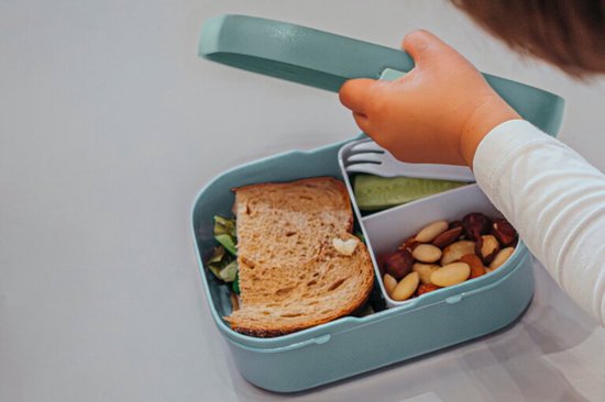 Un enfant ferme une boîte à lunch vert d'eau avec compartiment bento, remplie de pain, concombre et noix.