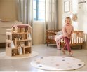 Houten poppenhuis met roze dak in een kinderkamer, naast een ledikantje. Een meisje met knuffel staat ernaast.