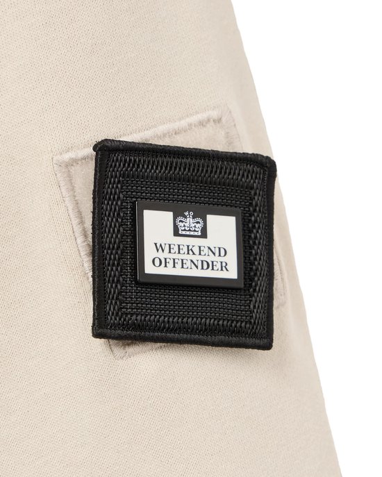 Detail van een beige trui met een zwart label met 'WEEKEND OFFENDER' erop.