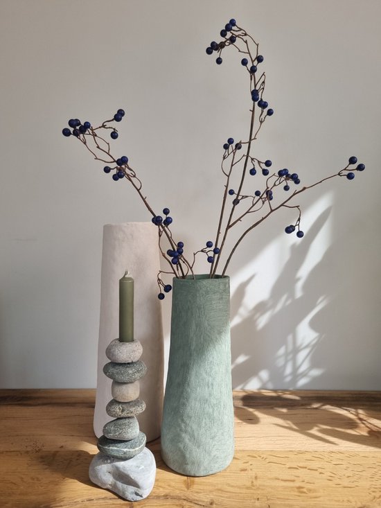 Lichtroze vaas met steenachtig oppervlak naast een groene vaas met takken met blauwe bessen en een stapel grijze stenen met groene kaars.
