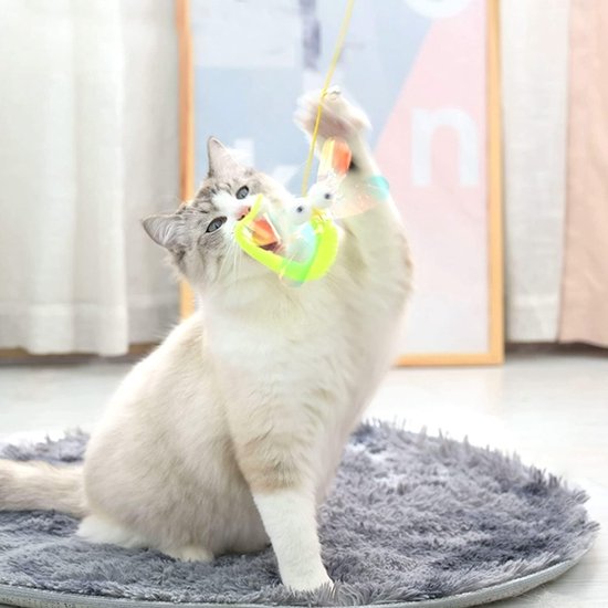 Een witte kat met grijze vlekken springt naar een libelle-vormig kattenspeeltje met glanzende vleugels en grote ogen, dat aan een koord hangt.