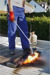 Man met rode handschoenen en blauw werkpak gebruikt brander op zwarte dakrol met vlammen.