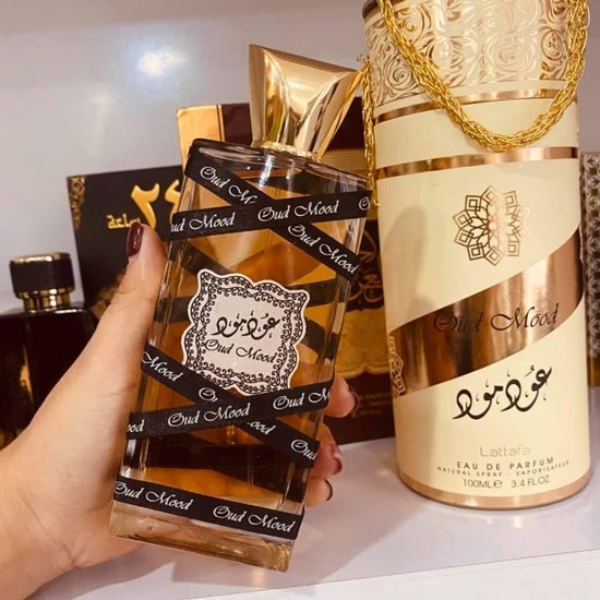 Hand houdt Oud Mood parfumfles met gouden dop, omwikkeld met zwart lint met "Oud Mood" erop. Naast een cilinderverpakking.