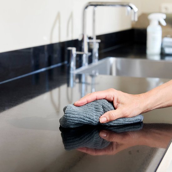 Une main avec un chiffon en microfibre gris nettoie un plan de travail noir.