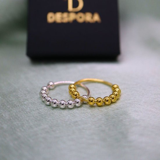 Twee ringen, één goudkleurig en één zilverkleurig, met elkaar verbonden door kleine balletjes. Zwarte verpakking op de achtergrond.