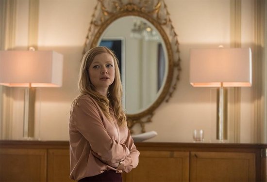 Een vrouw met blond haar in een roze satijnen blouse en rode rok, staand met de armen over elkaar.