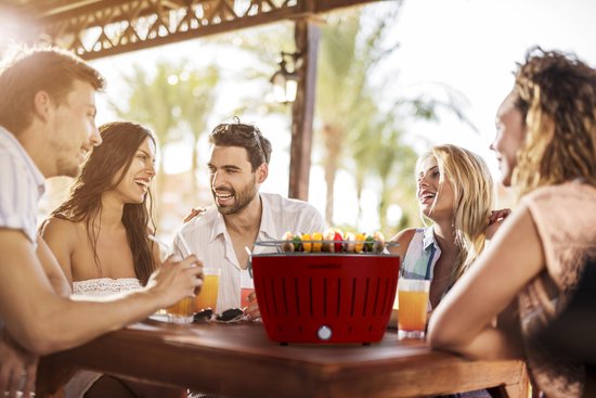 Groep vrienden geniet van eten rond een rode LotusGrill aan een tafel.