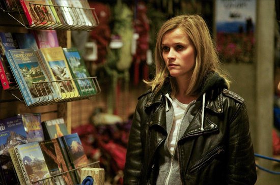 Reese Witherspoon aux cheveux blonds, porte une veste en cuir noire sur un sweat gris et un t-shirt blanc. Elle se tient dans un magasin près d'un présentoir de livres.